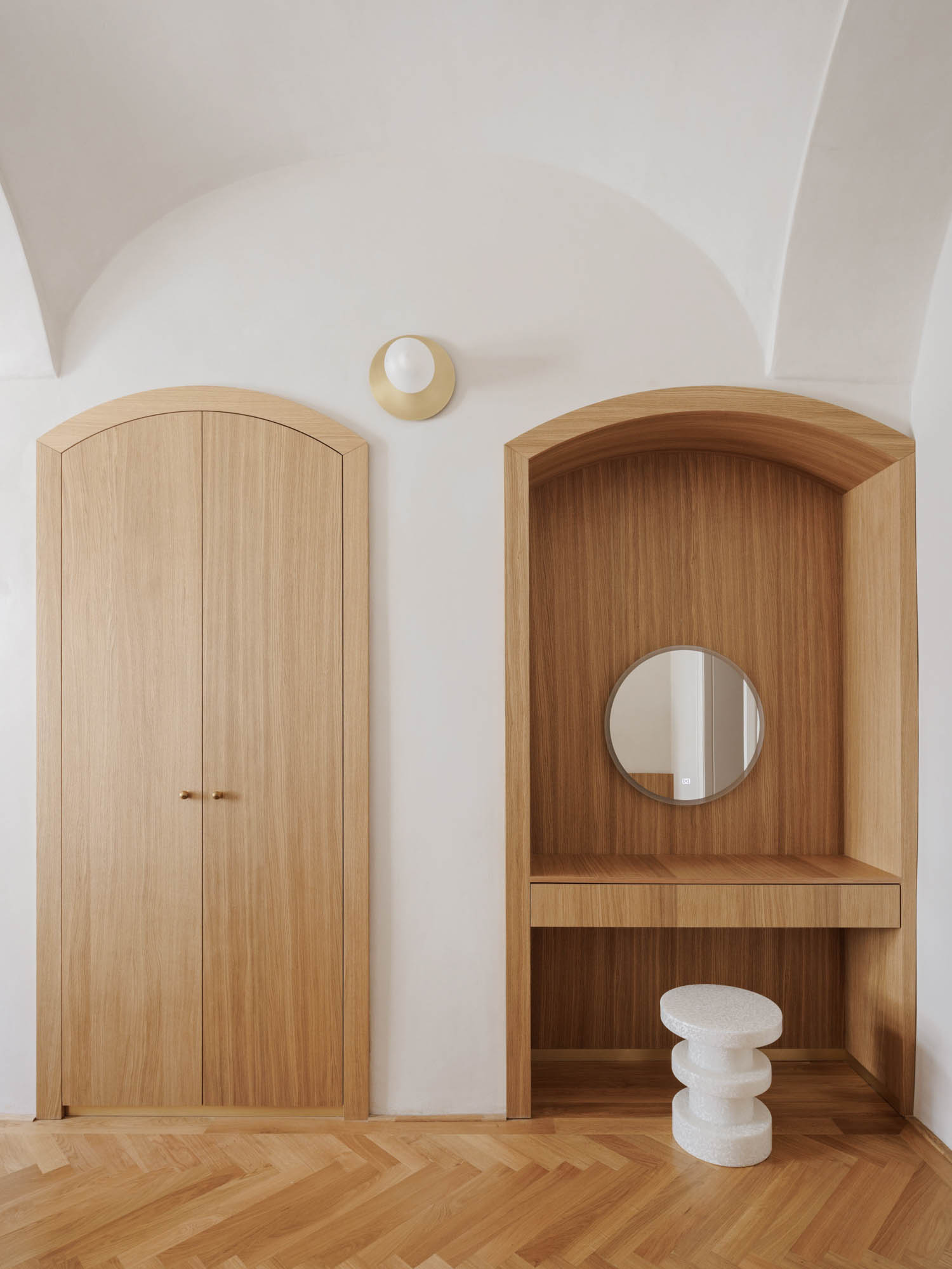 An arched wooden closet next to a matching arched vanity nook with a circular mirror and a small white stool.