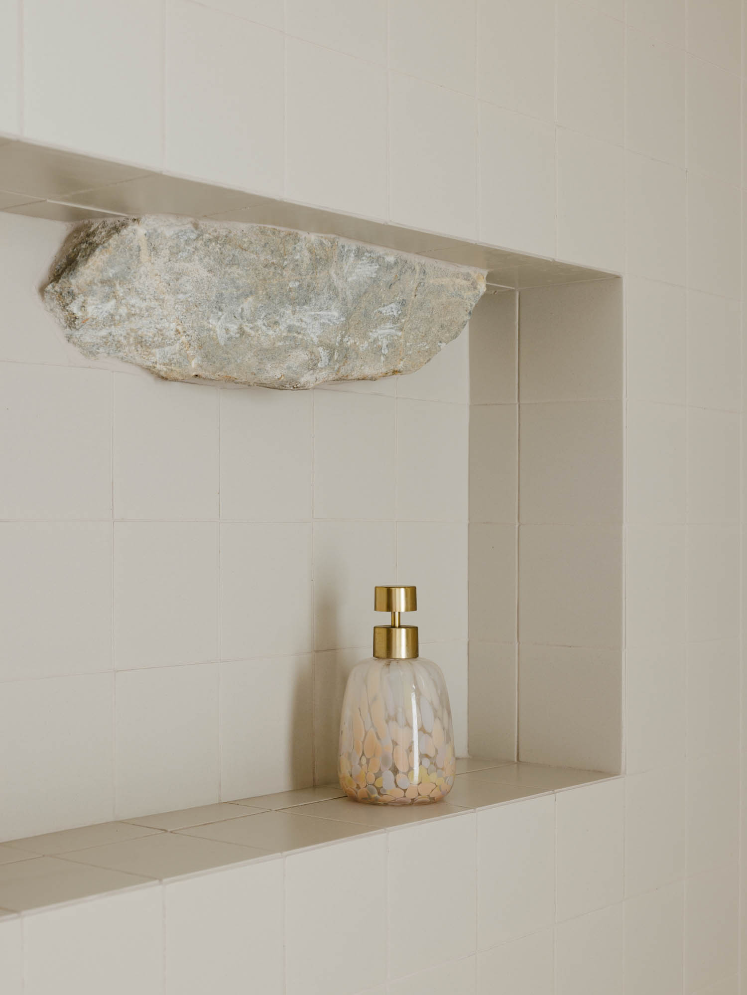 A soap dispenser with a gold pump sits in a tiled shower niche containing a jagged piece of raw stone.