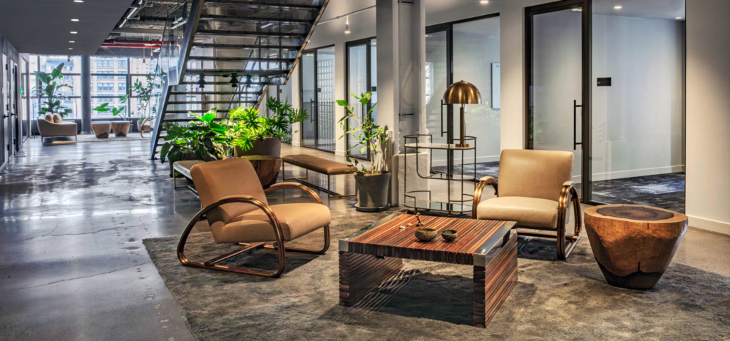 A modern office lobby area with two tan leather chairs, a wooden slat coffee table, a natural wood side table, and several potted plants.