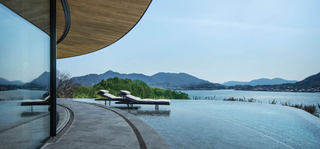 Two lounge chairs positioned in an infinity pool overlooking a bay and mountains.