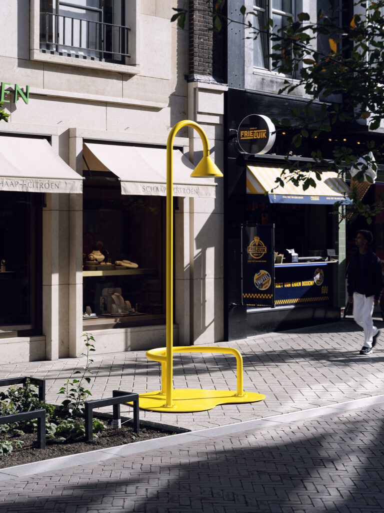 A bright yellow sculpture shaped like a giant spilled drink or drop on a city sidewalk in front of storefronts.
