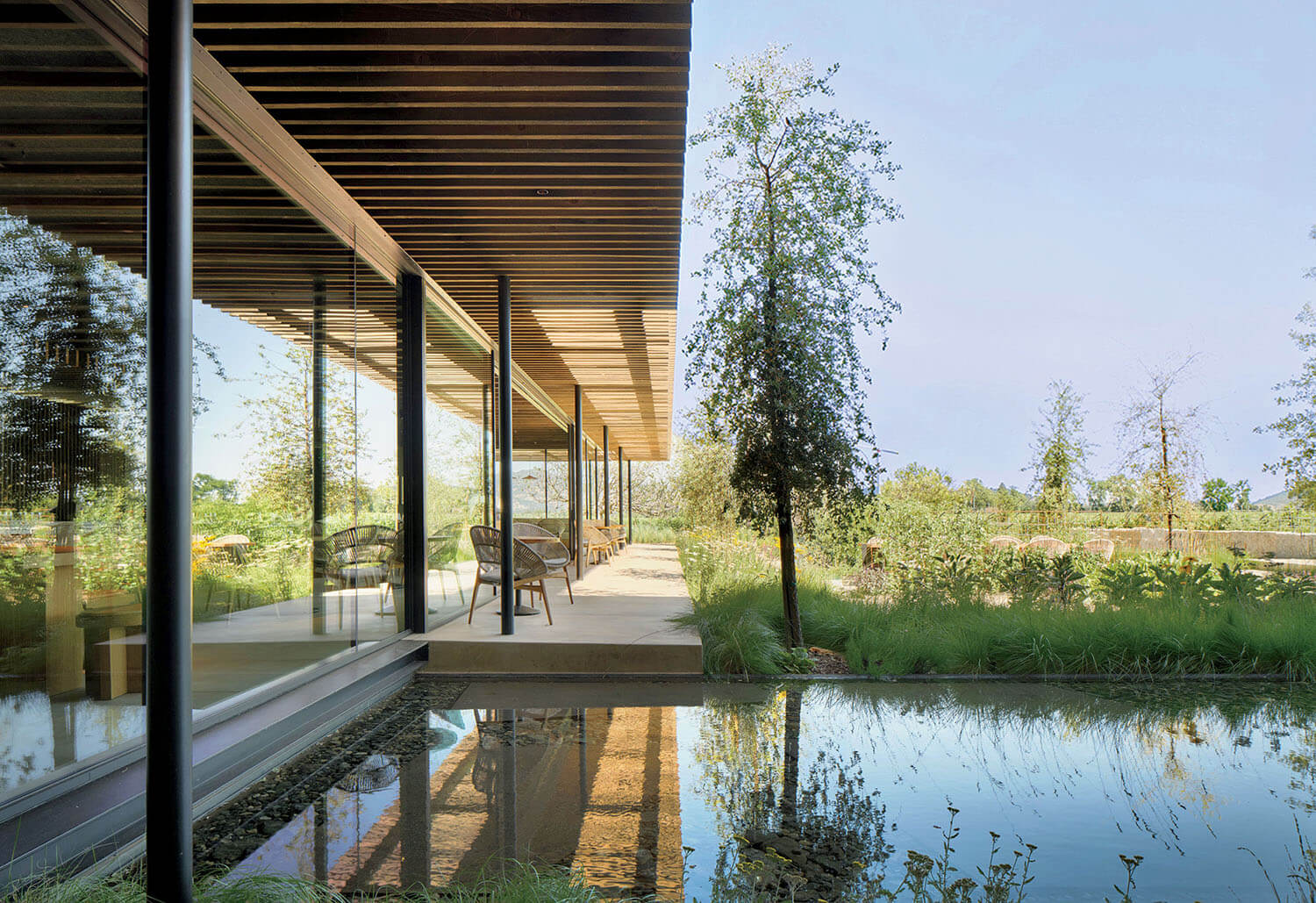 Modern building with floor-to-ceiling glass walls and an outdoor patio, overlooking a garden and a reflective pond.
