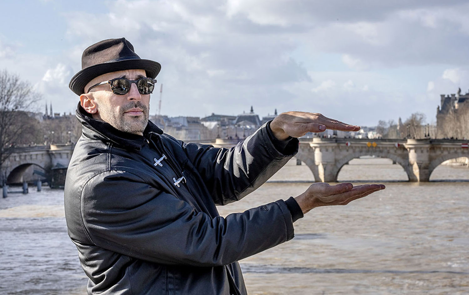 A man wearing a hat and sunglasses gestures as if holding a small object between his hands, with a river and stone bridge in the background.