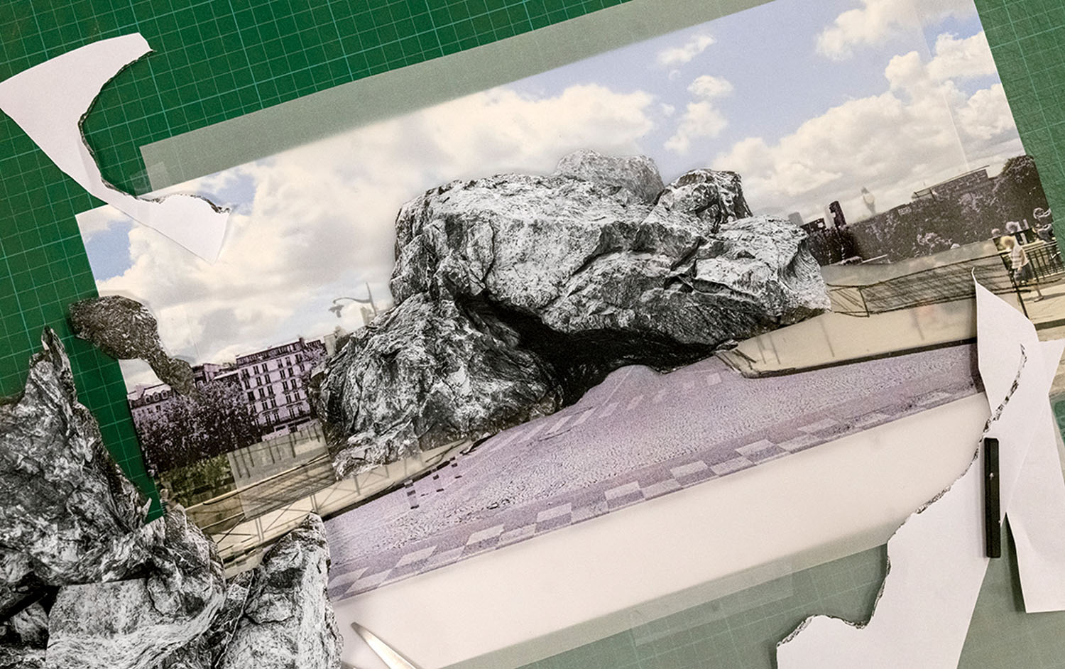 A collage featuring a large, textured rock cutout layered over a printed street scene, placed on a green cutting mat with scattered paper scraps.