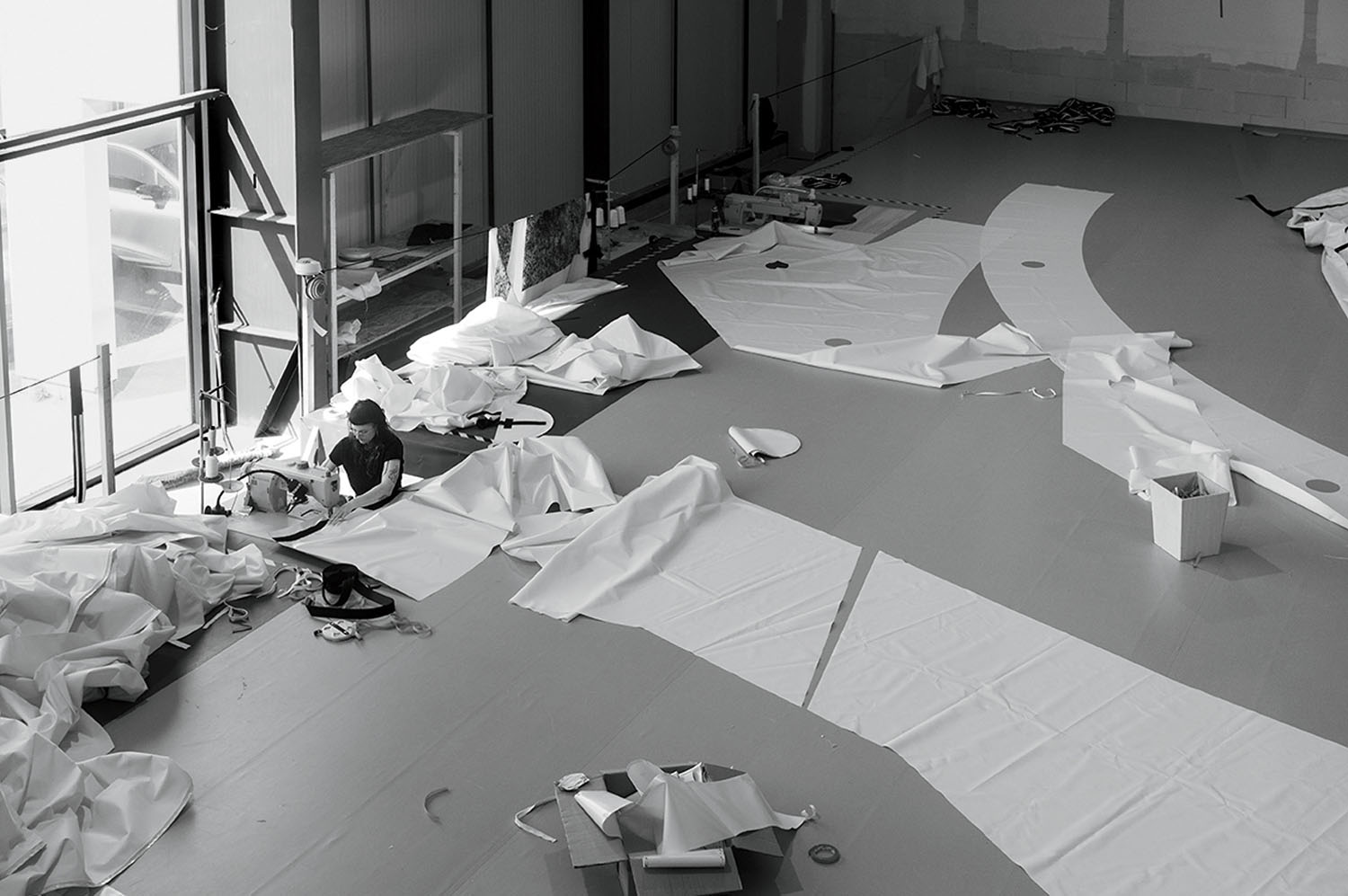 A person using a sewing machine to join large white fabric panels on the floor of a workshop.
