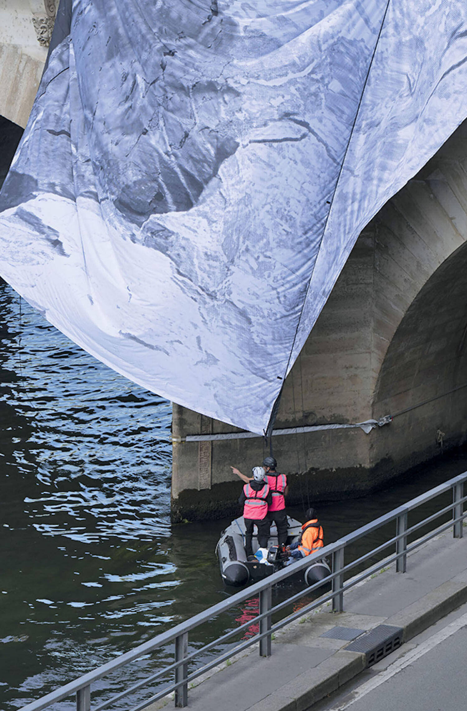 Three people in a small motorboat under a stone bridge, holding up a large canvas printed with a black-and-white mountain landscape.
