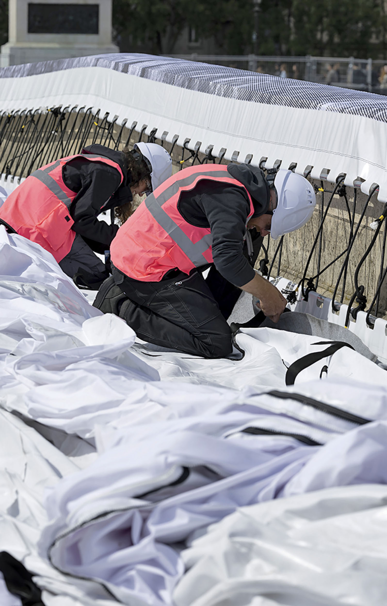 Two construction workers in white hard hats and pink safety vests kneeling to secure large white fabric coverings.