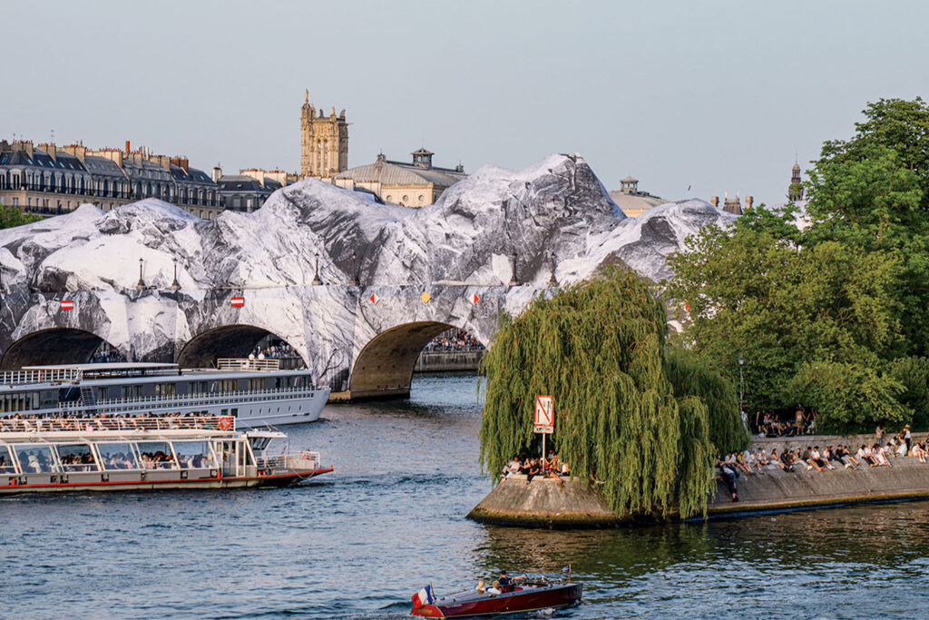 Pont Neuf bridge in Paris wrapped in a black and white mountain-like covering, with tour boats on the Seine and people gathered on a nearby riverbank.