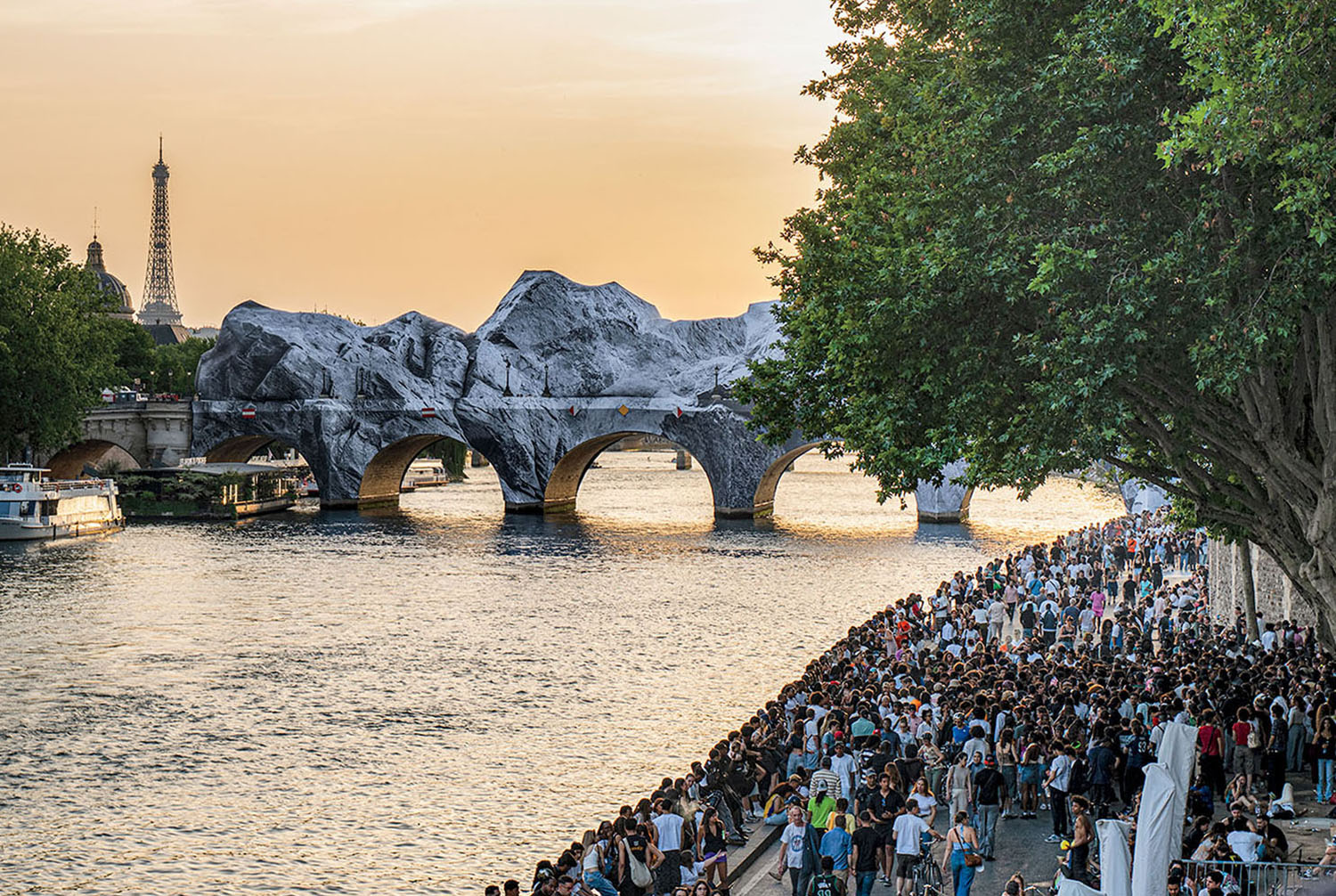A large crowd gathered along the banks of the Seine river in Paris, with the Pont Neuf bridge wrapped in fabric and the Eiffel Tower in the distance.