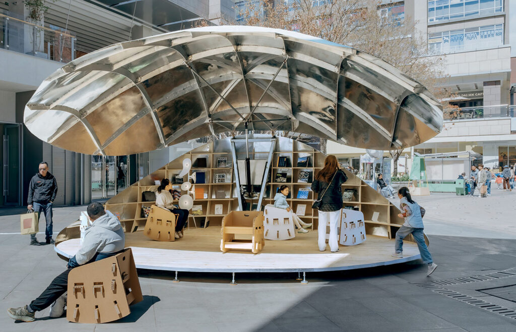 People use a circular outdoor reading pavilion featuring a large reflective canopy and modular cardboard furniture.