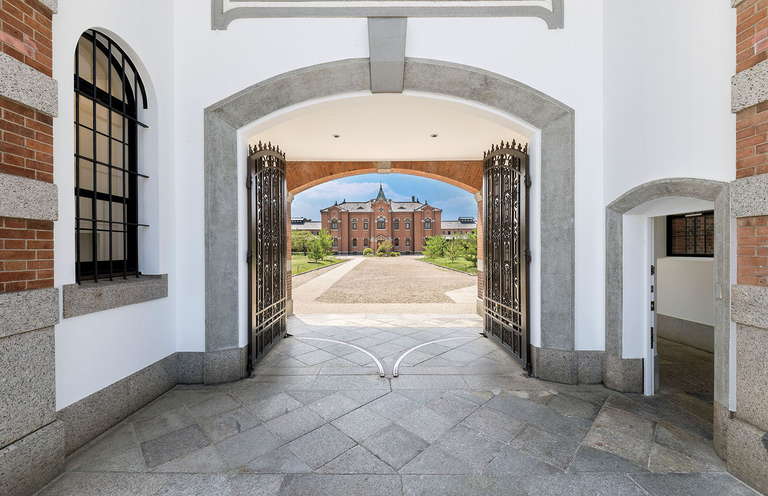 A view through an open arched stone gateway onto a gravel courtyard leading to a large, multi-story brick building.