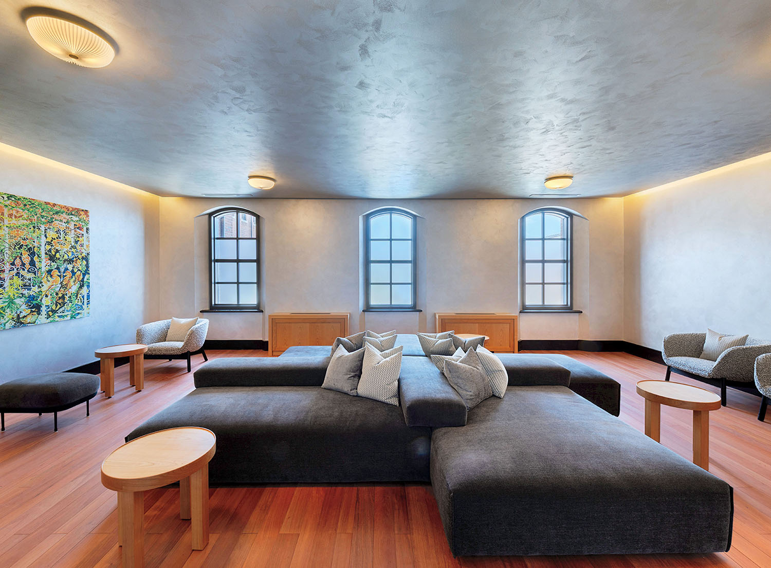 A lounge area with a large central sectional sofa, small wooden tables, and two accent chairs near three arched windows.