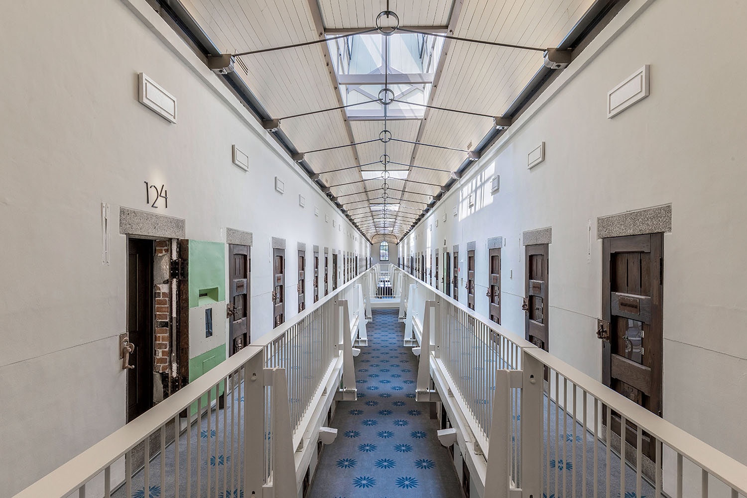 A long, narrow prison corridor with two levels of cells behind white railings and a blue patterned floor.