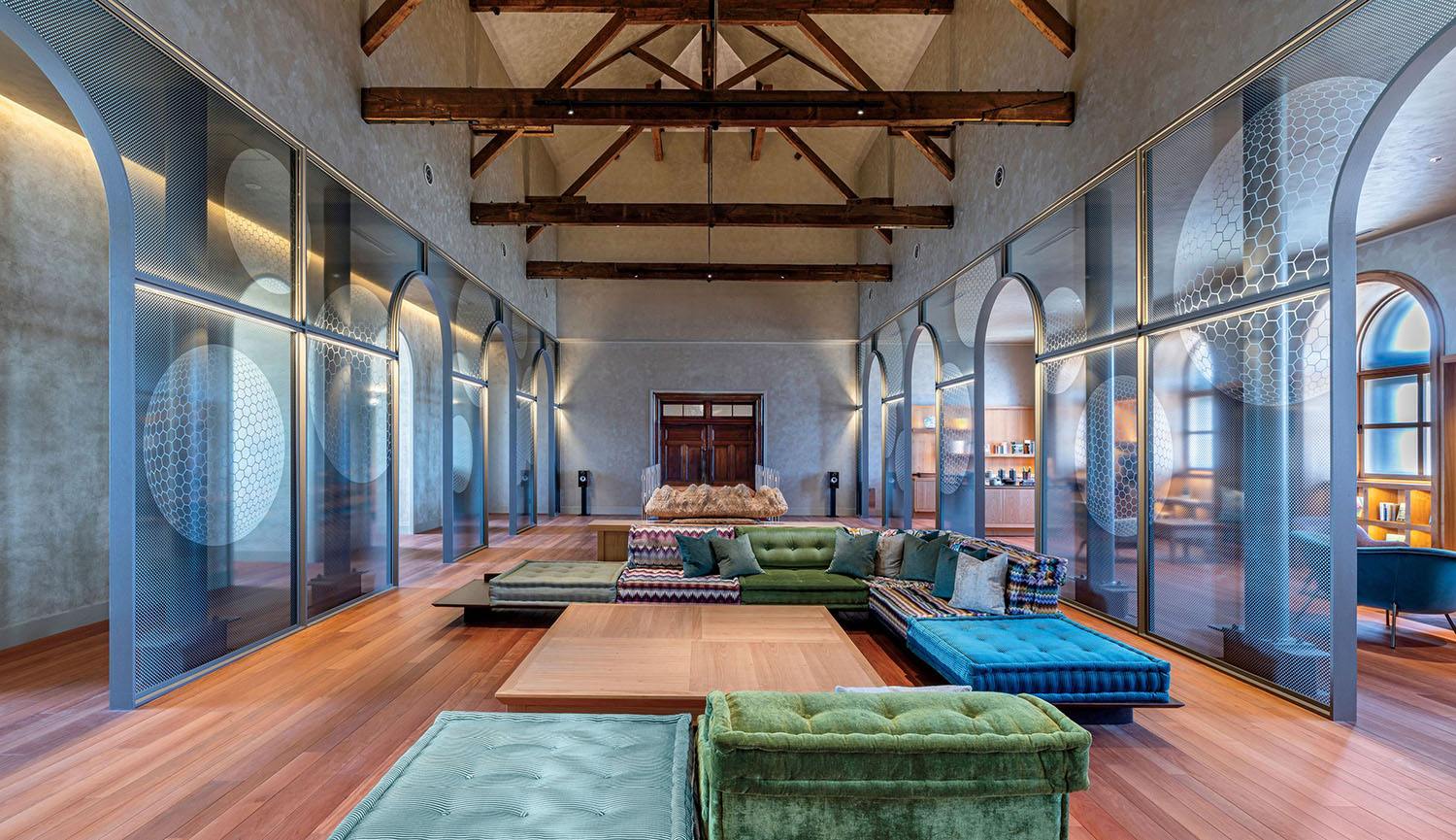 A large, open room with exposed wooden ceiling beams, modular green and blue floor seating, and glass partitions etched with circular geometric patterns.