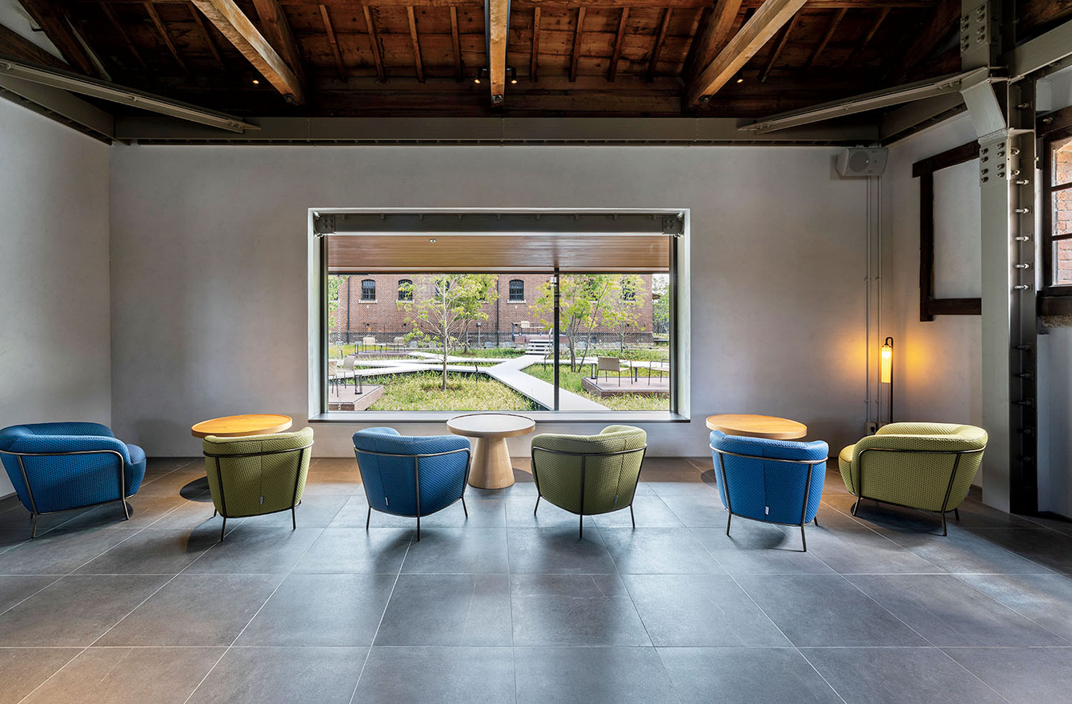 Six armchairs in blue and green arranged in pairs around small tables, facing a large window overlooking a landscaped courtyard.