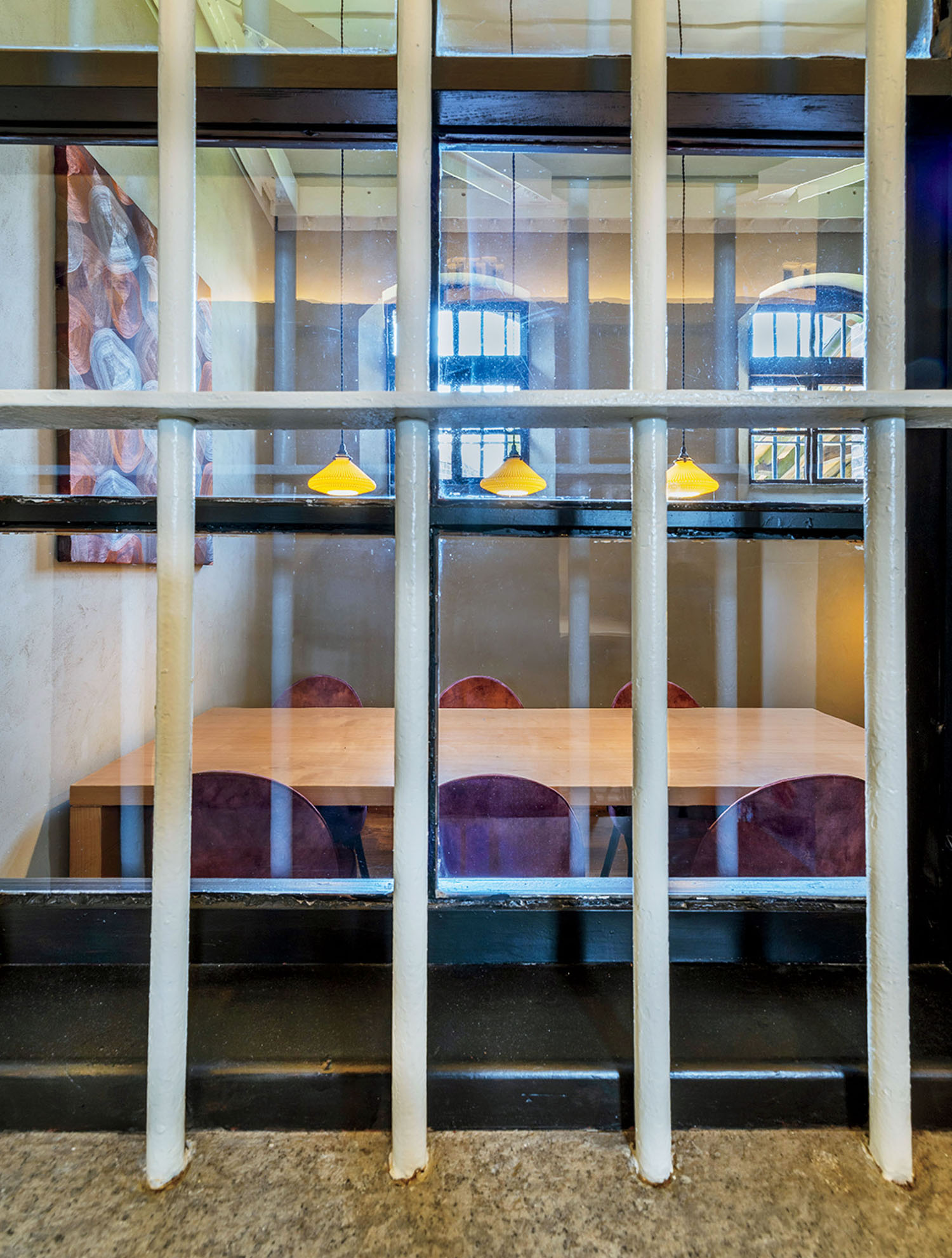 A wooden table with purple chairs seen through a window with white vertical security bars and three yellow pendant lights.