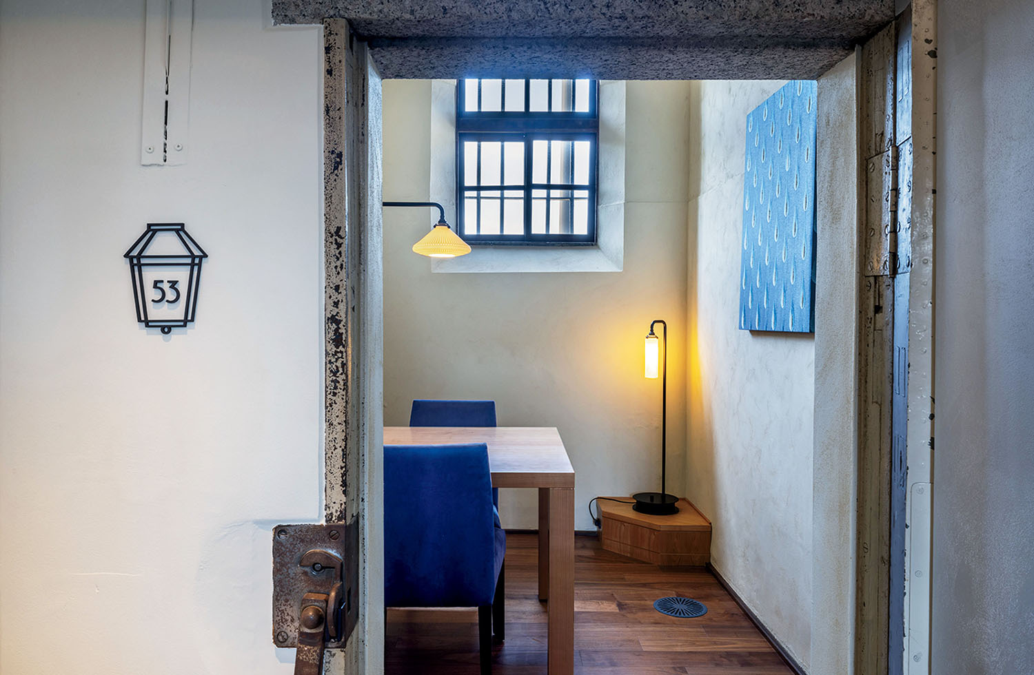 A view through a stone doorway into a small room with a wooden table, two blue chairs, a window with metal bars, and two lamps. A sign marked “53” is on the wall to the left.