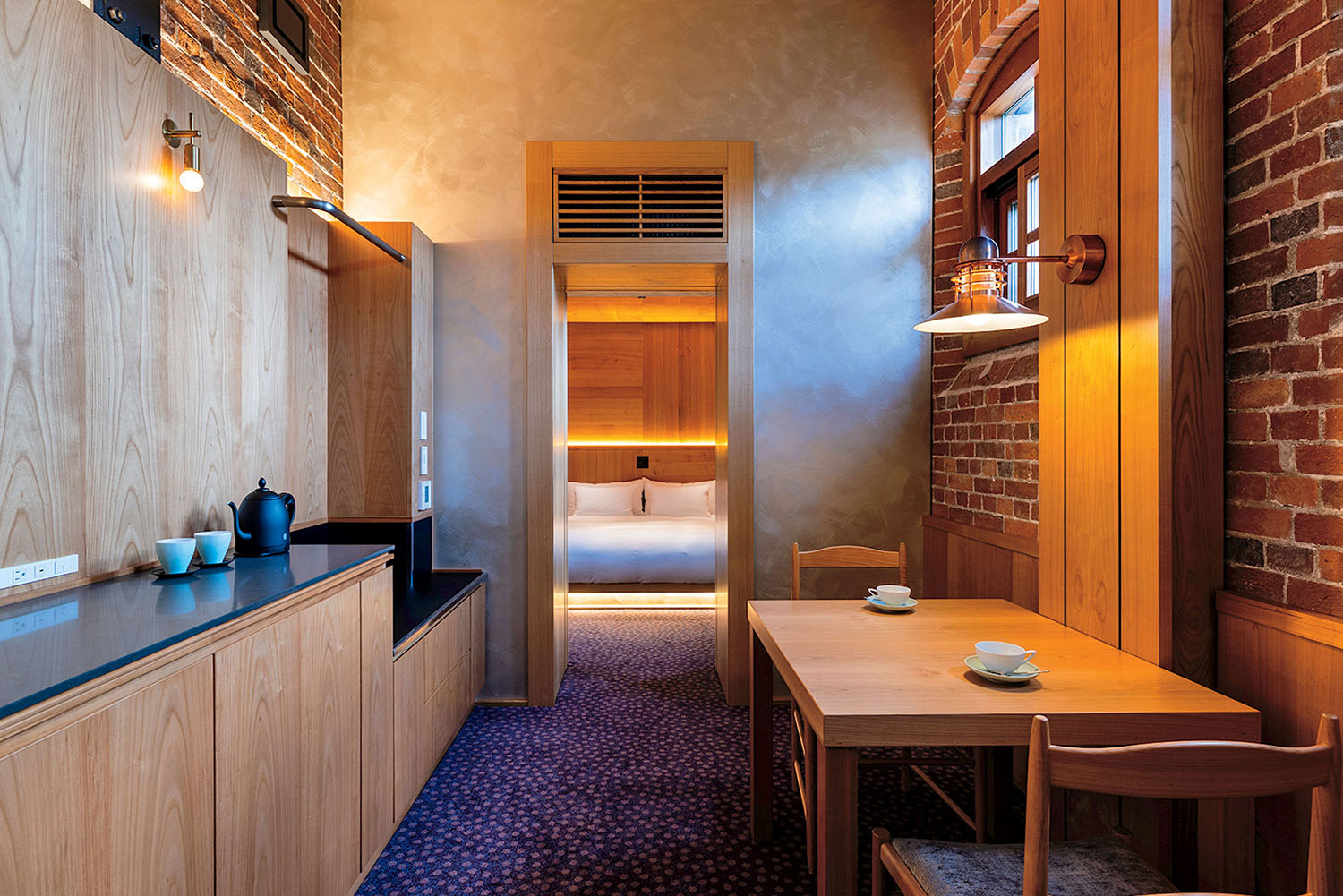 A hotel room featuring a kitchenette with a kettle, a wooden dining table, and a view into a bedroom with a bed.