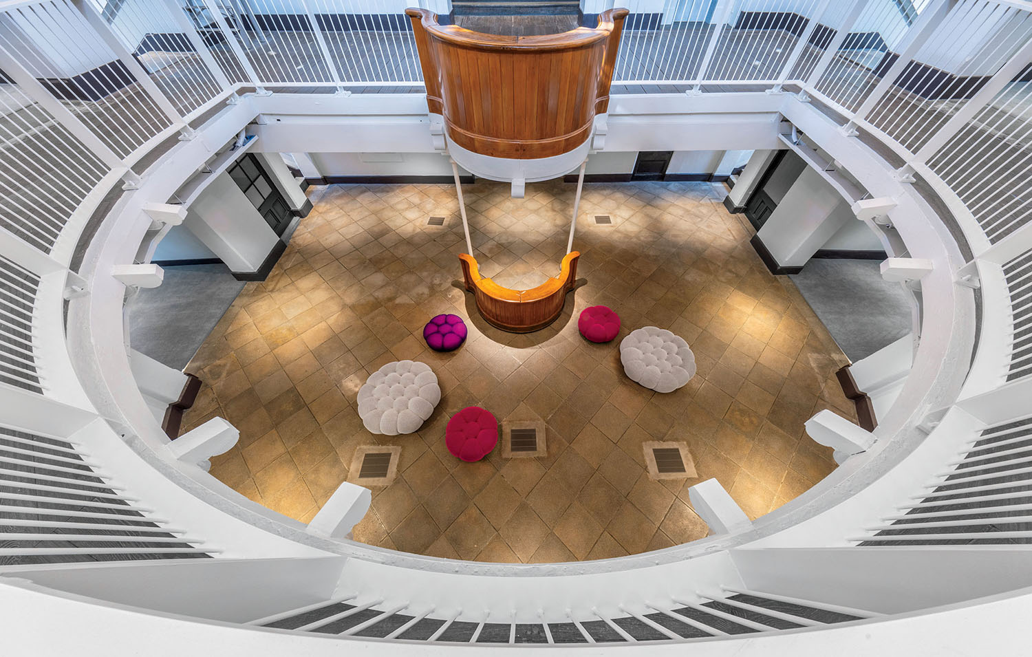 High-angle view of a circular building interior with a wooden balcony overlooking a ground floor lobby containing round purple, pink, and white tufted ottomans.