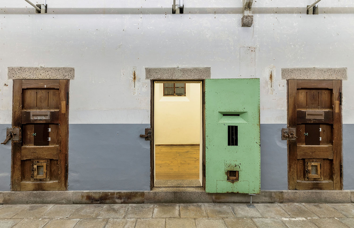 A row of three prison cell doors, one of which is open, set into a grey and white wall.