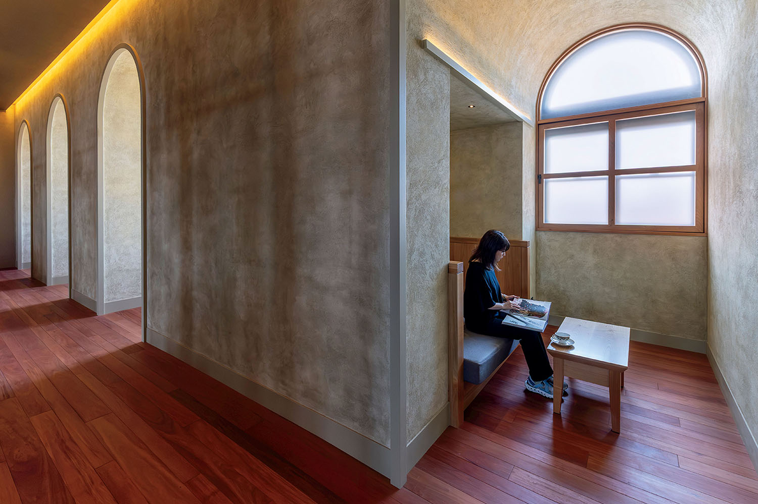 A person sits on a bench reading in a recessed alcove with an arched window, next to a small wooden table and a cup of coffee.