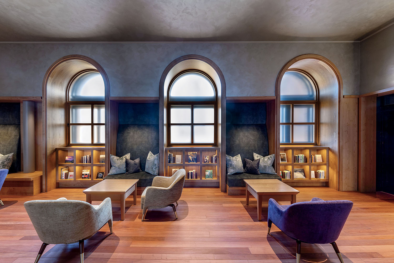 Lounge area with three arched windows, built-in shelving, cushioned seating nooks, and four armchairs arranged around two wooden coffee tables.