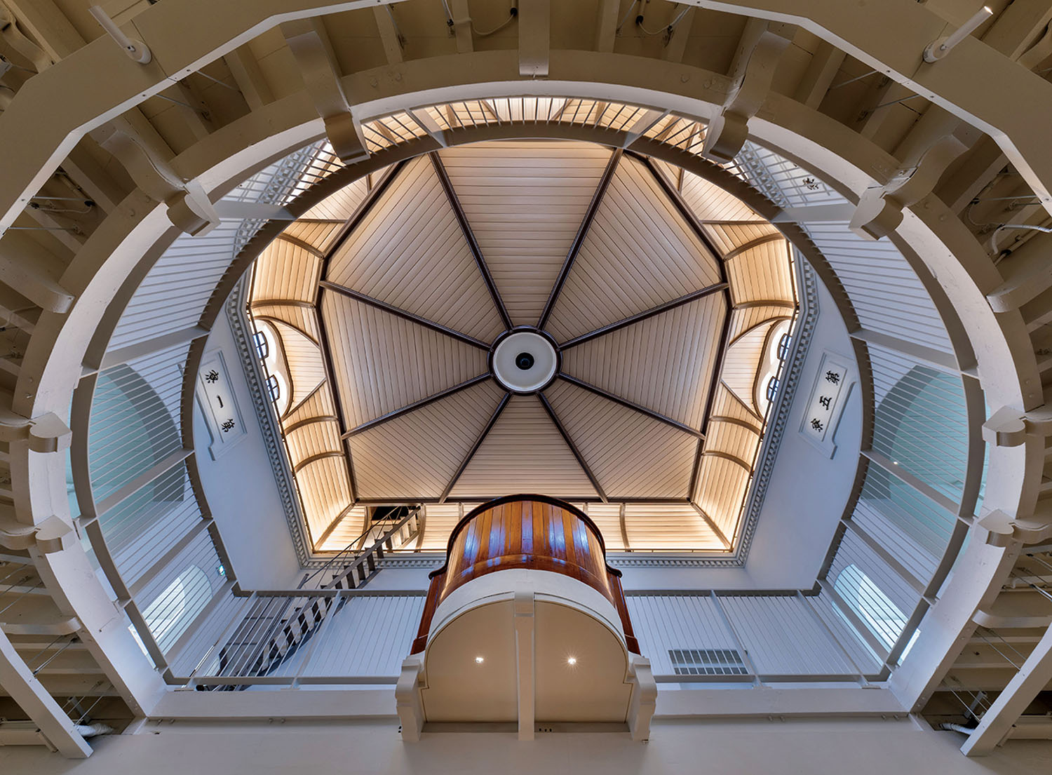 Looking upward at the vaulted, octagonal ceiling of an interior space with a central curved wooden balcony and a metal ladder.