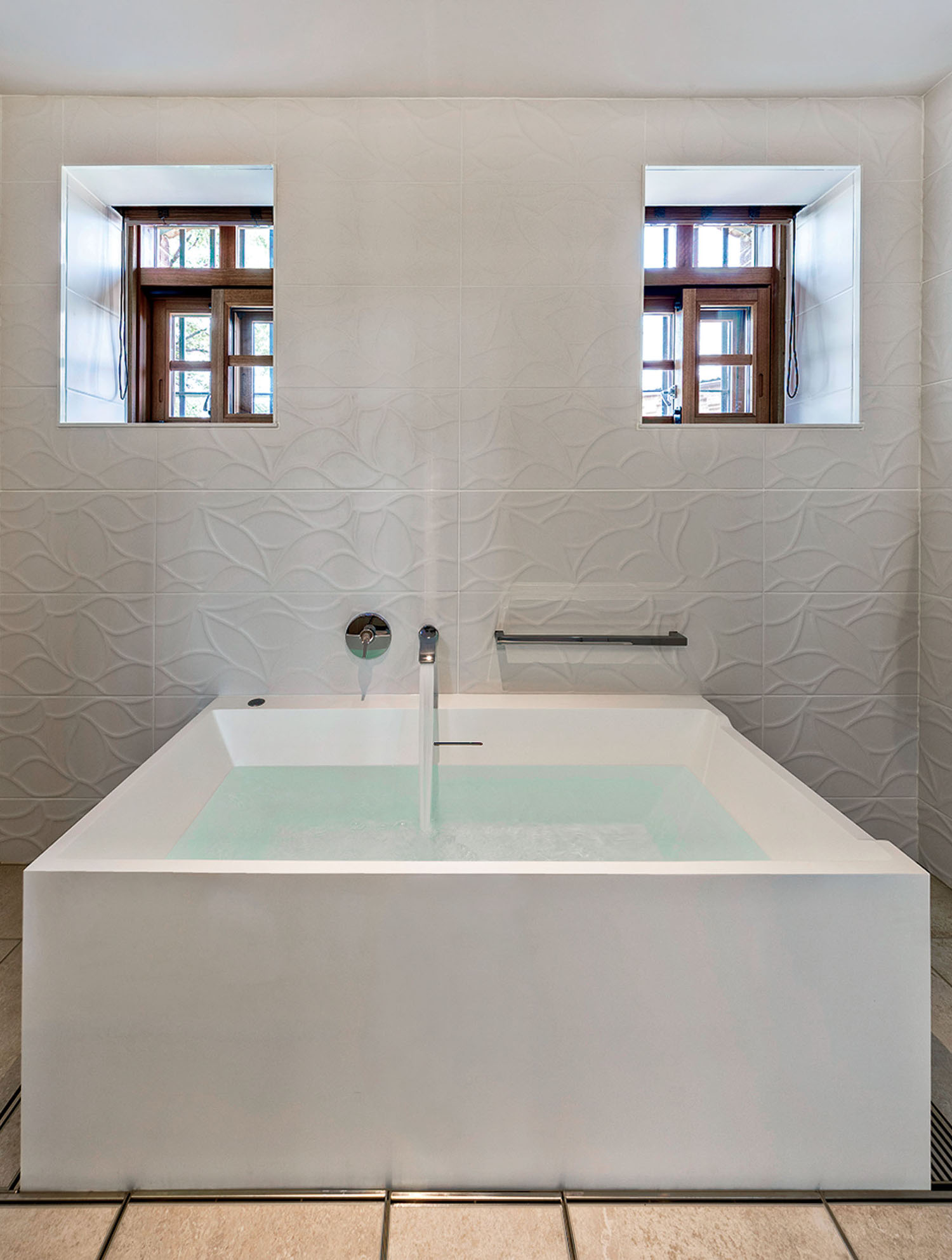 A square white bathtub filled with water, centered in front of two small windows set into a textured tiled wall.