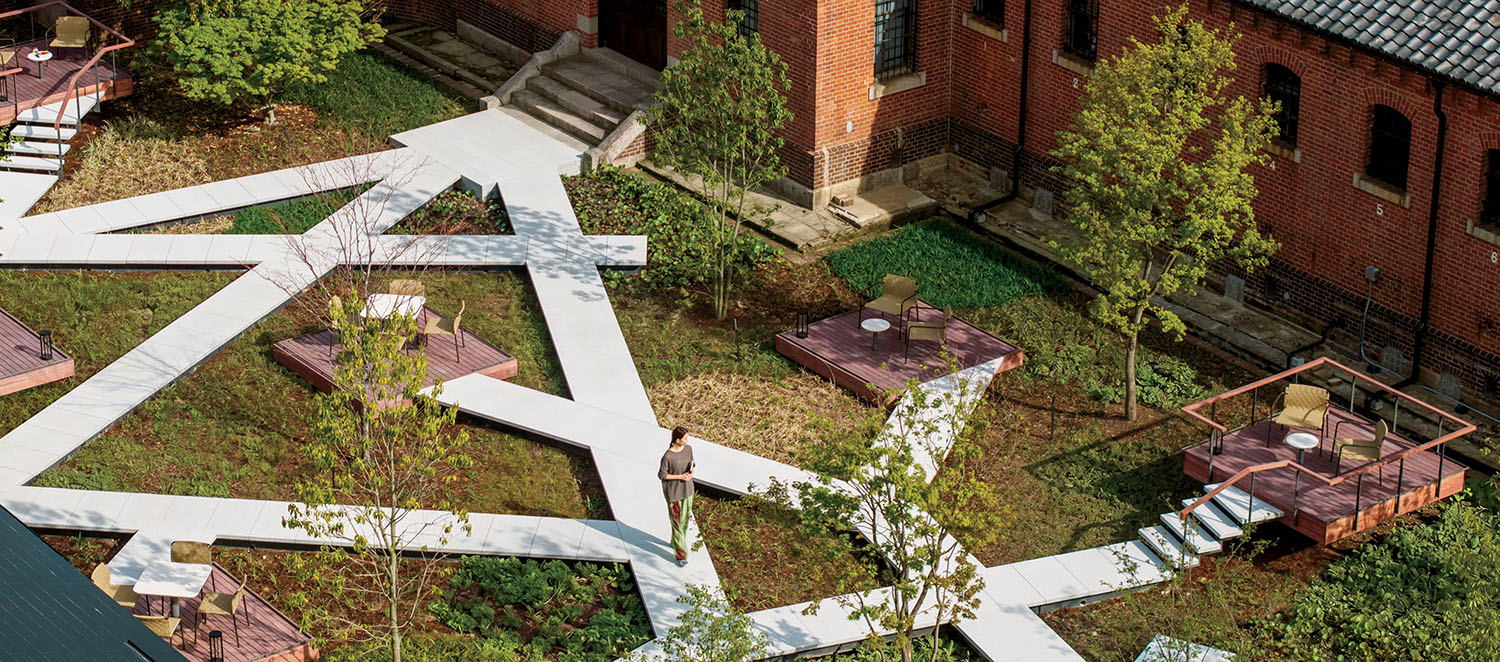 Aerial view of a garden with geometric white paths, wooden seating platforms, and a brick building.