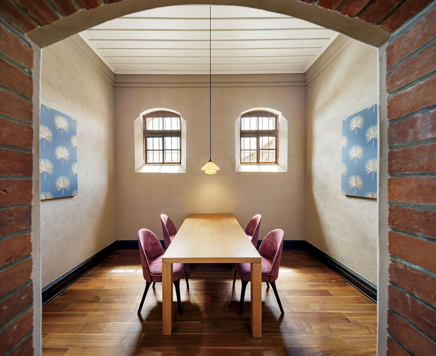 A dining room framed by a brick archway, featuring a wooden table with four pink upholstered chairs, two windows, and two blue wall prints.