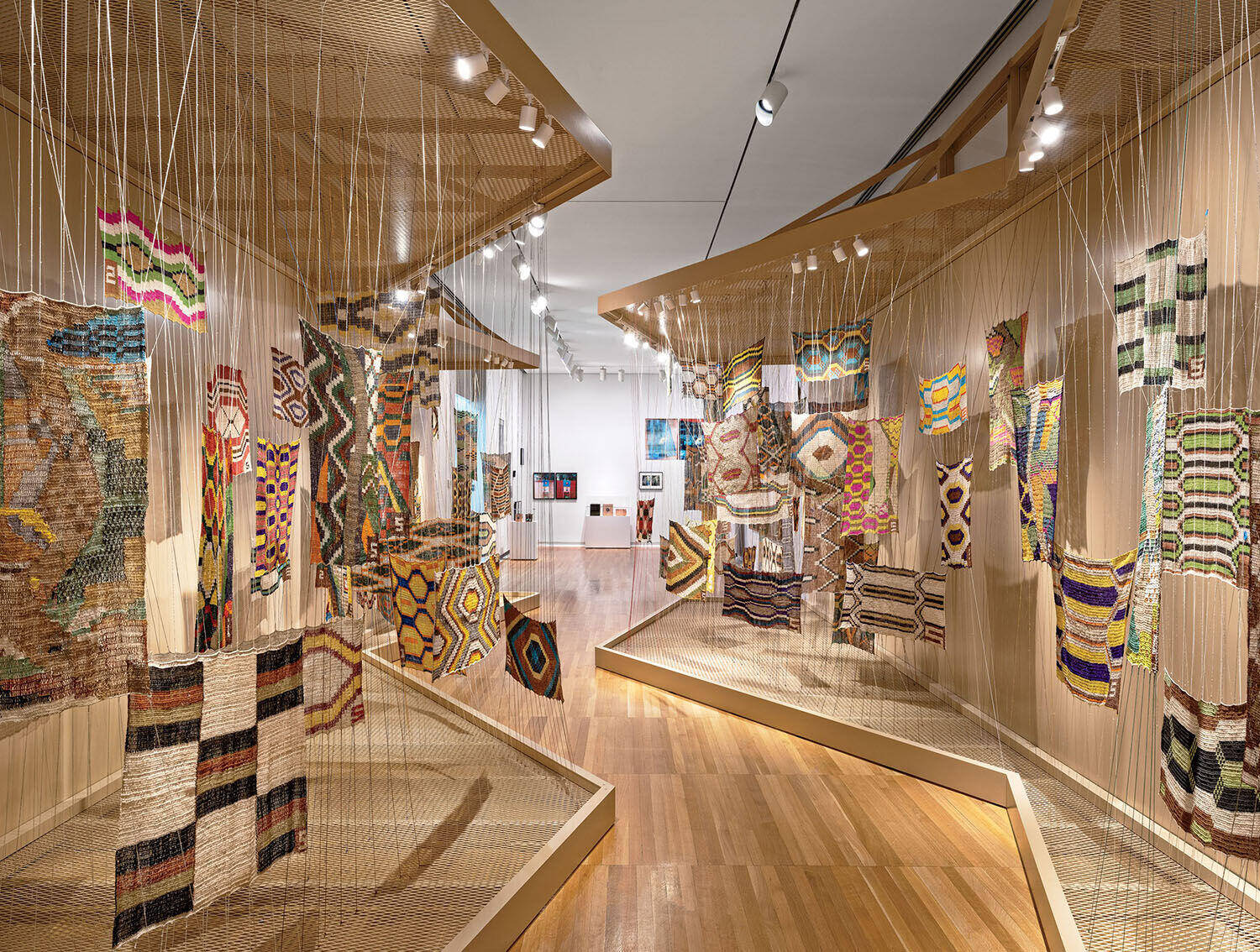 An art installation featuring many colorful, rectangular woven textile panels suspended from thin wires in a gallery space.