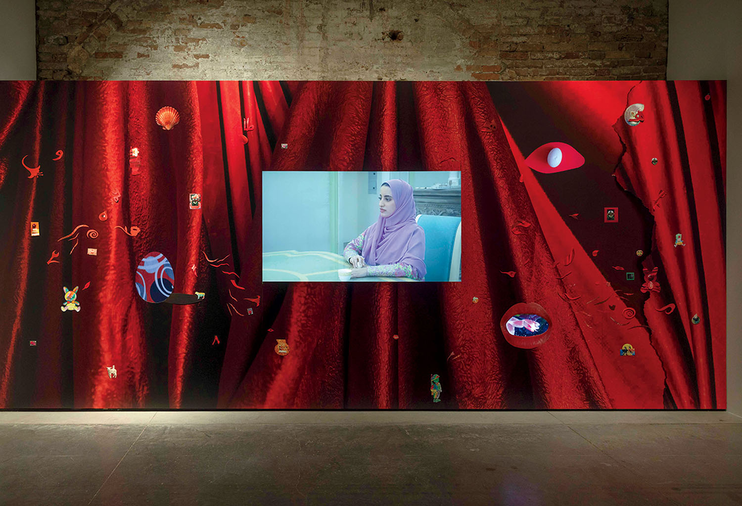 A screen showing a woman in a lavender hijab sits in the center of a large, red-curtained digital display decorated with small, floating icons.
