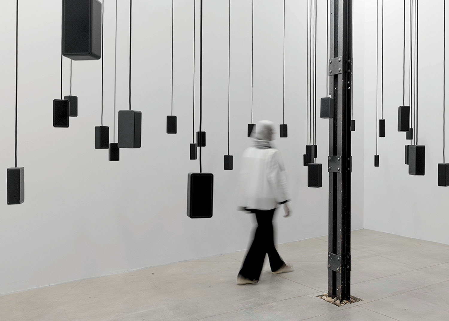 A person in a white top and black trousers walks through a room filled with black rectangular speakers hanging at various heights from the ceiling by black cords.