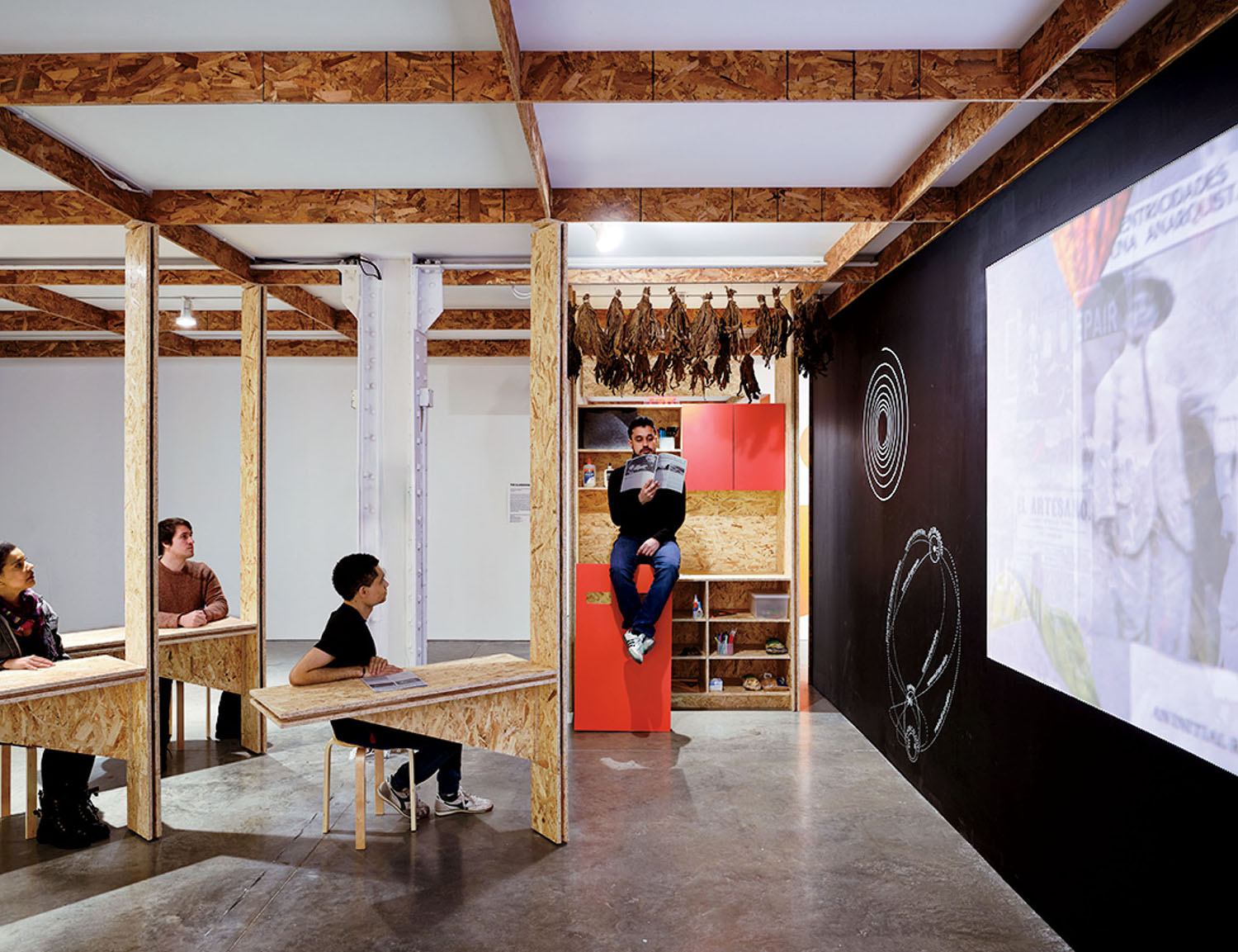 Three people sit at desks facing a large wall projection, while a person sits on a cabinet reading a book in an industrial space with wood-framed partitions.