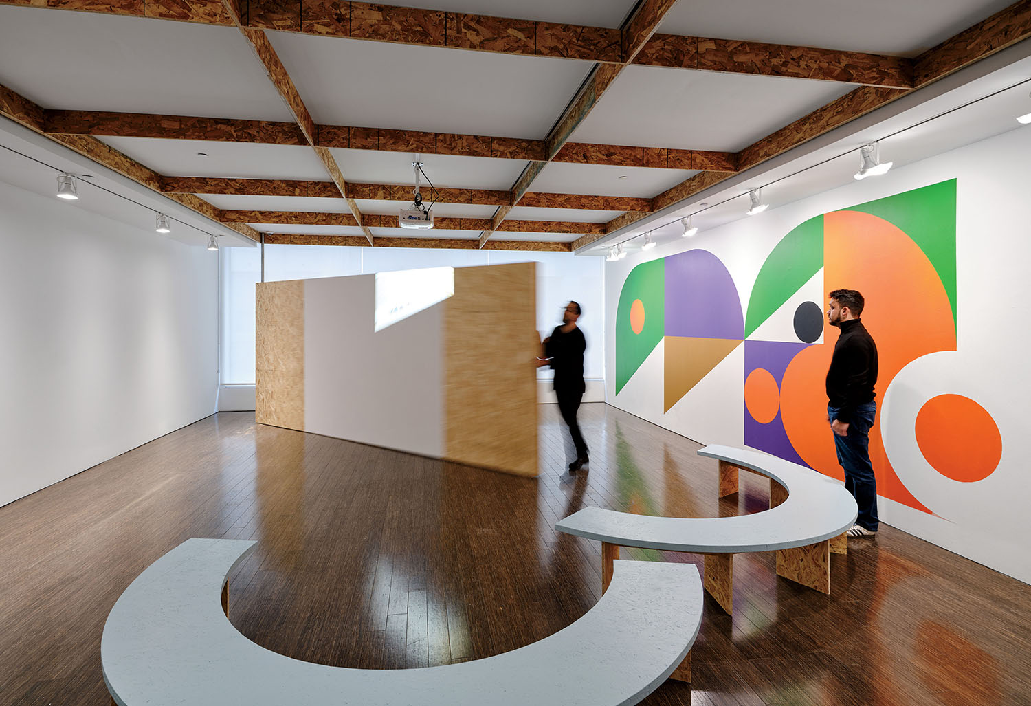 A person walks past a large particle-board partition in an art gallery, while another person stands by a large, colorful geometric mural.