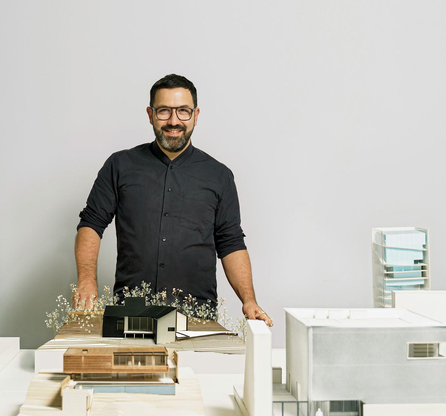 A bearded man in a dark button-down shirt stands behind a table featuring several architectural scale models.