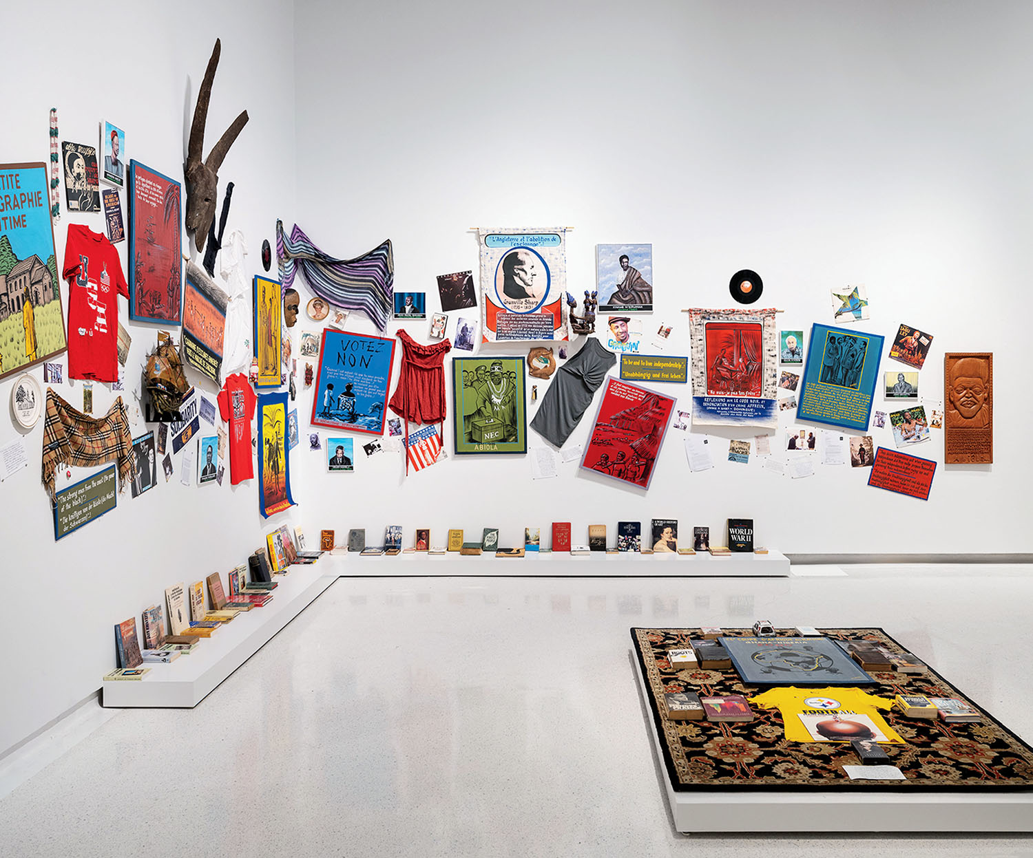 A gallery installation featuring a wide array of posters, clothing, books, and artifacts displayed on white walls and low shelves, with a patterned rug topped with additional objects on the floor.