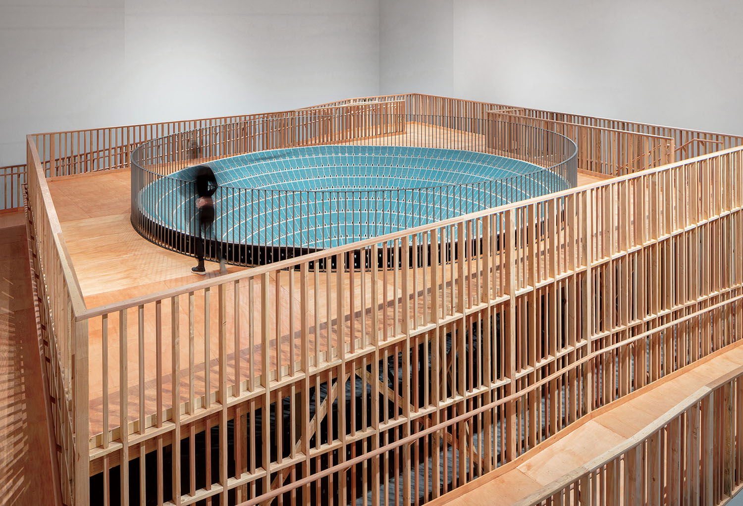 A person stands on a wooden platform overlooking a large, circular blue pool surrounded by a wooden railing.