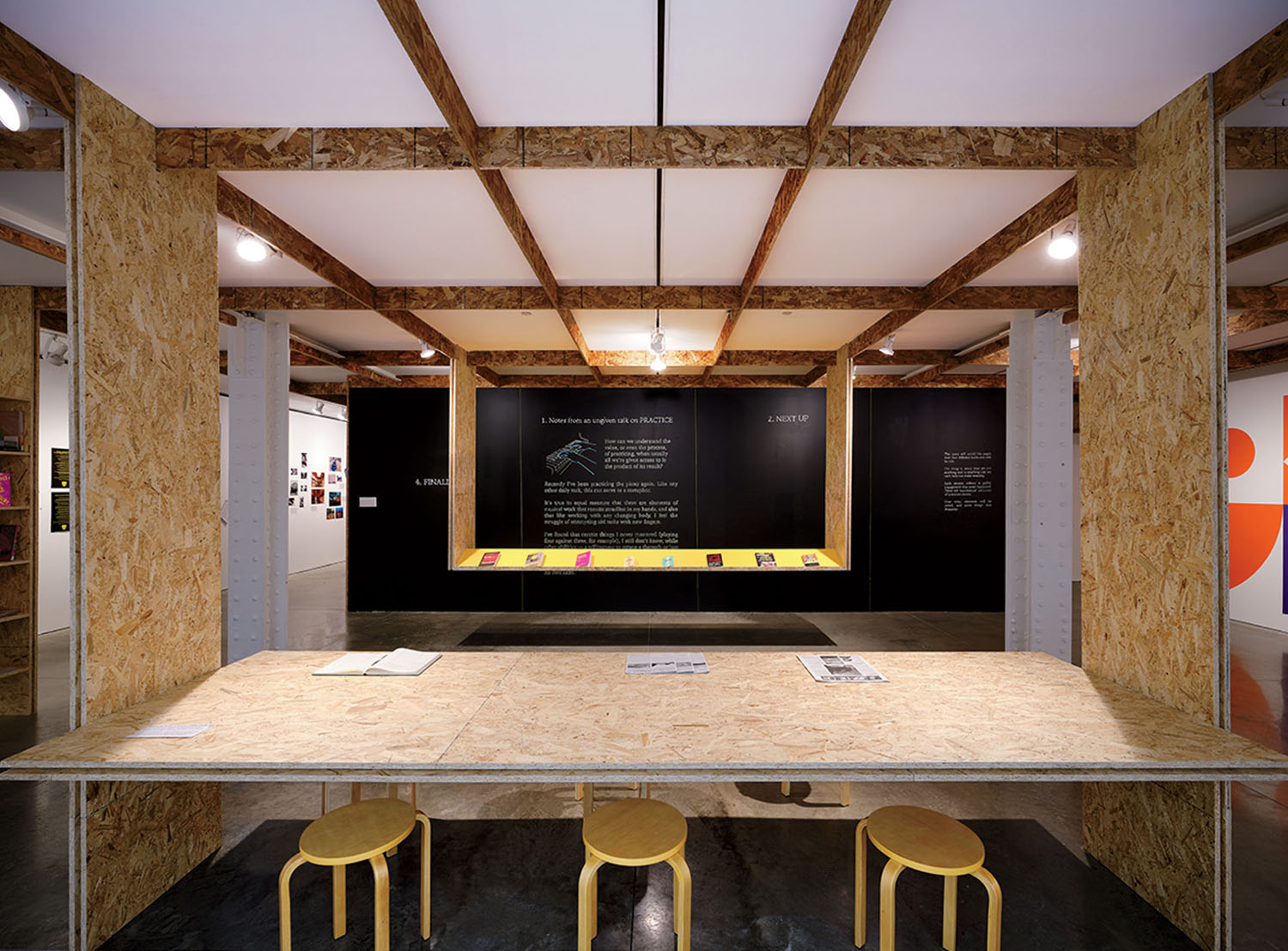 A wooden table with three stools faces a wall featuring a central display of small books and explanatory text under a wooden beam structure.
