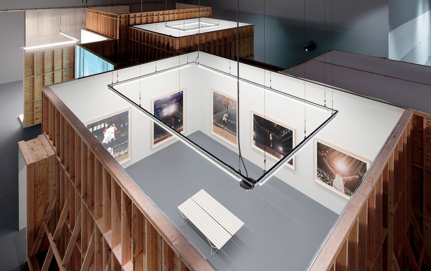 An open-roof gallery structure built from wooden studs, containing a white bench and framed sports photographs on the interior walls.