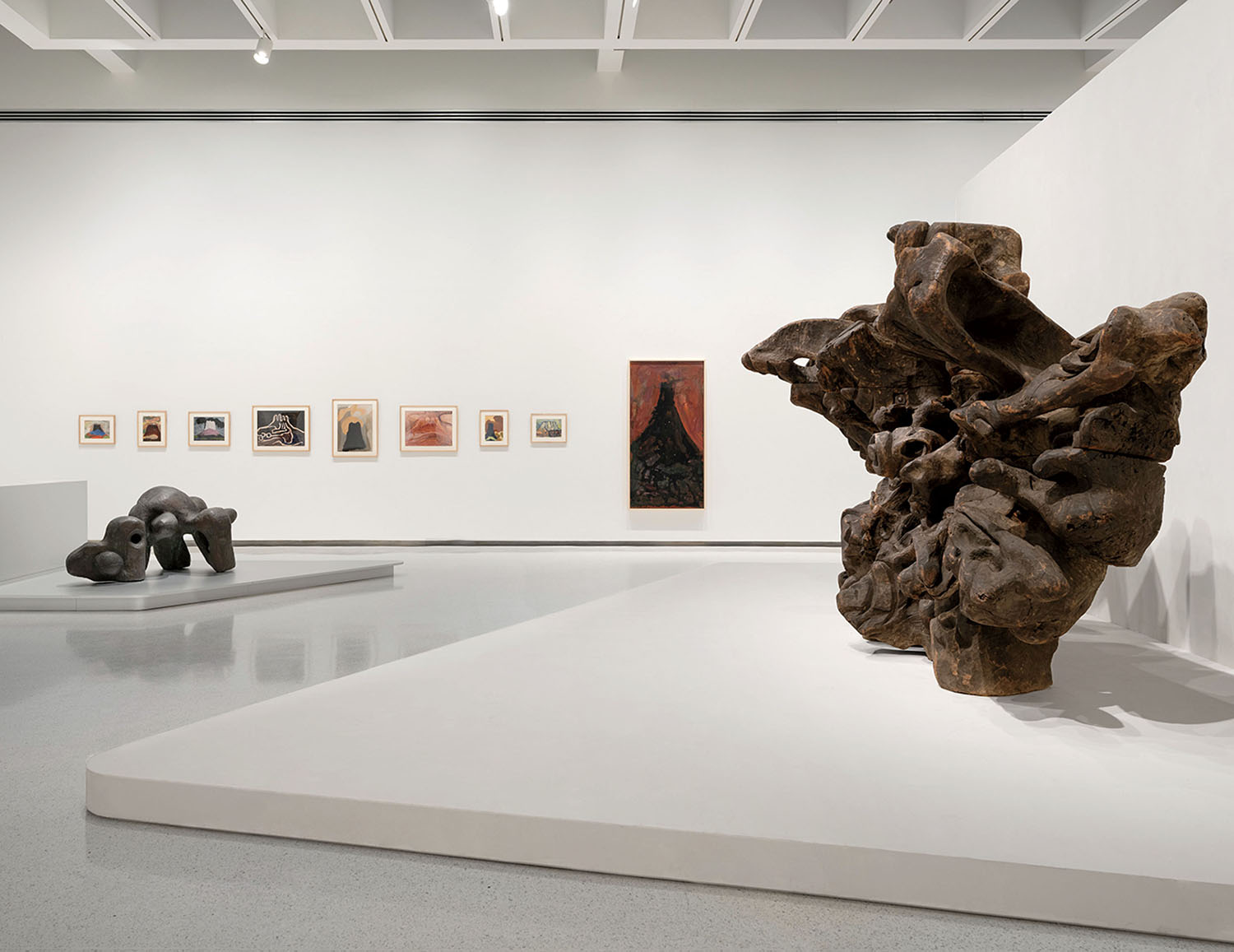 Gallery installation featuring two large, organic-shaped sculptures on white pedestals and a series of framed artworks on the wall.