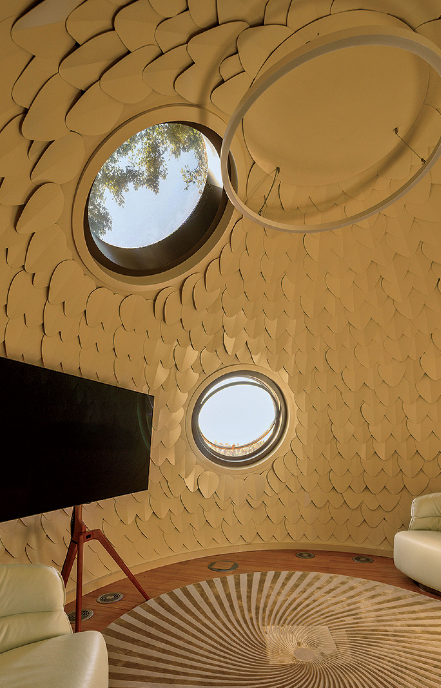 Interior of a dome-shaped room with scale-like wall paneling, two circular windows, a flat-screen TV on a tripod stand, and white armchairs on a patterned rug.