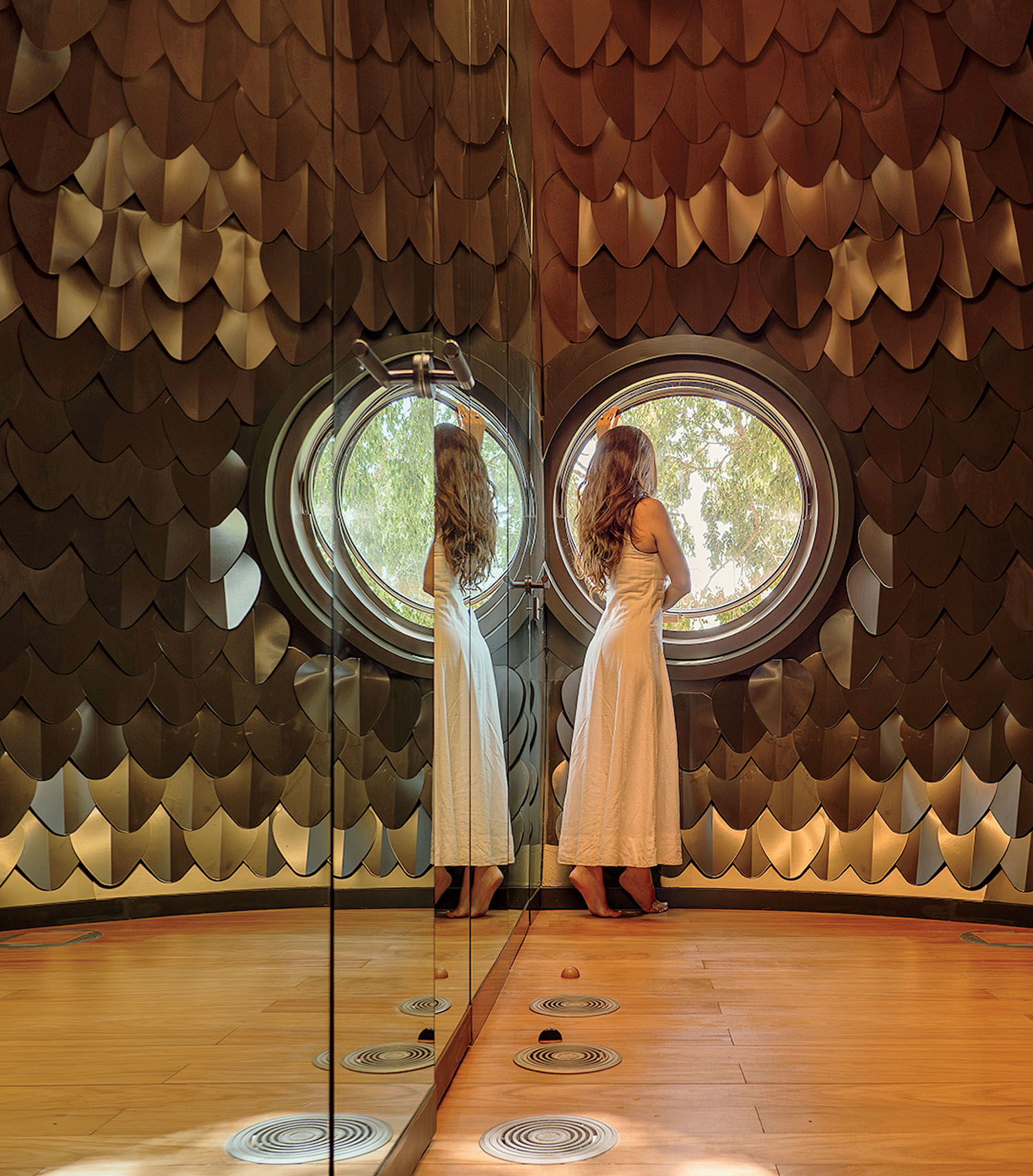A woman in a white dress stands looking out of a circular window in a room with walls covered in overlapping metal scales.