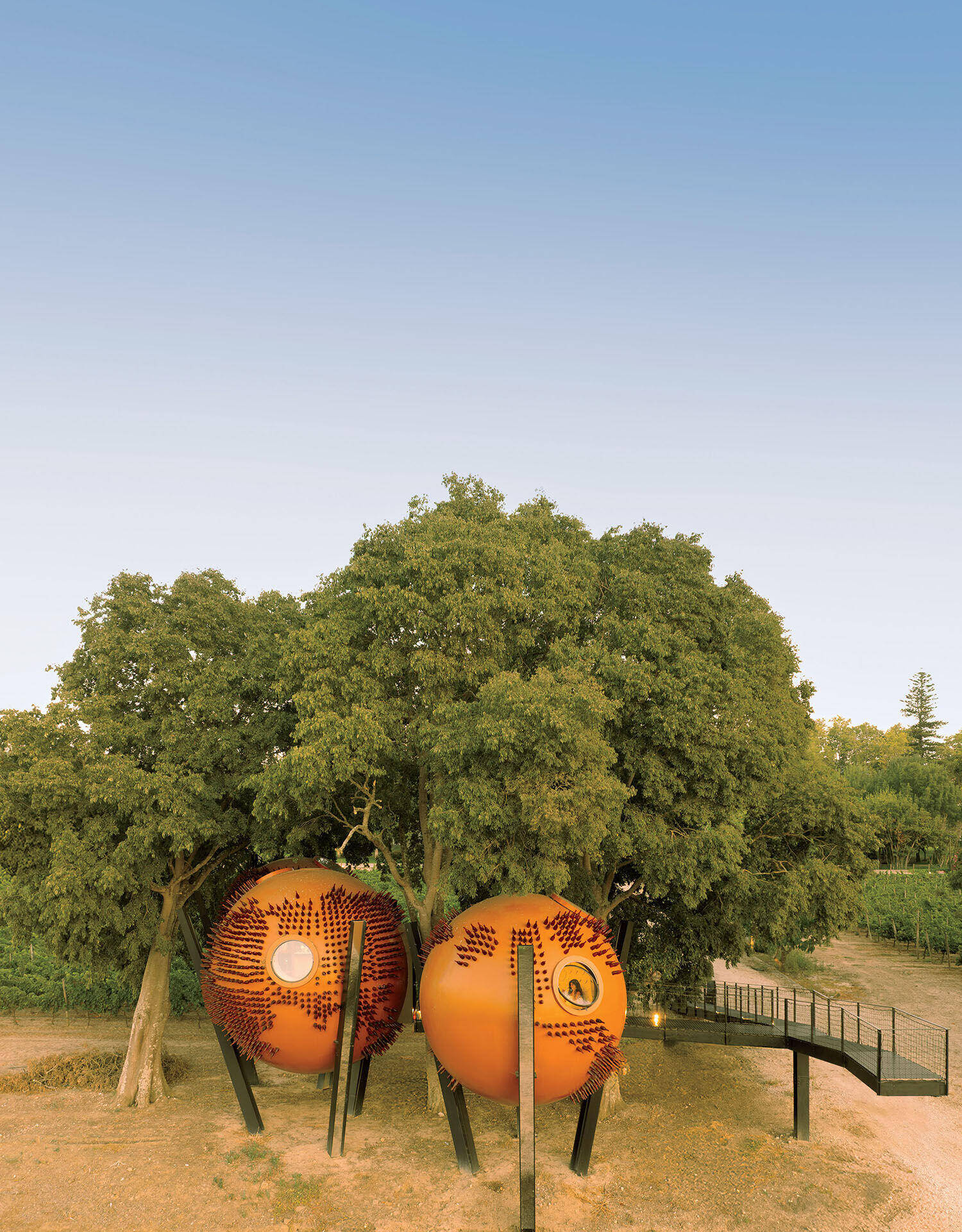 Two spherical orange structures with circular windows and textured surfaces elevated on poles beneath large trees, connected by a wooden walkway.