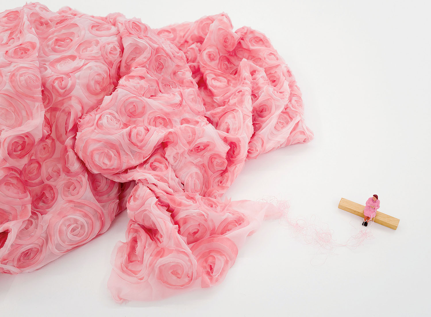A miniature figure sits on a wooden block, trailing a loose thread from a large pile of pink rose-patterned fabric.
