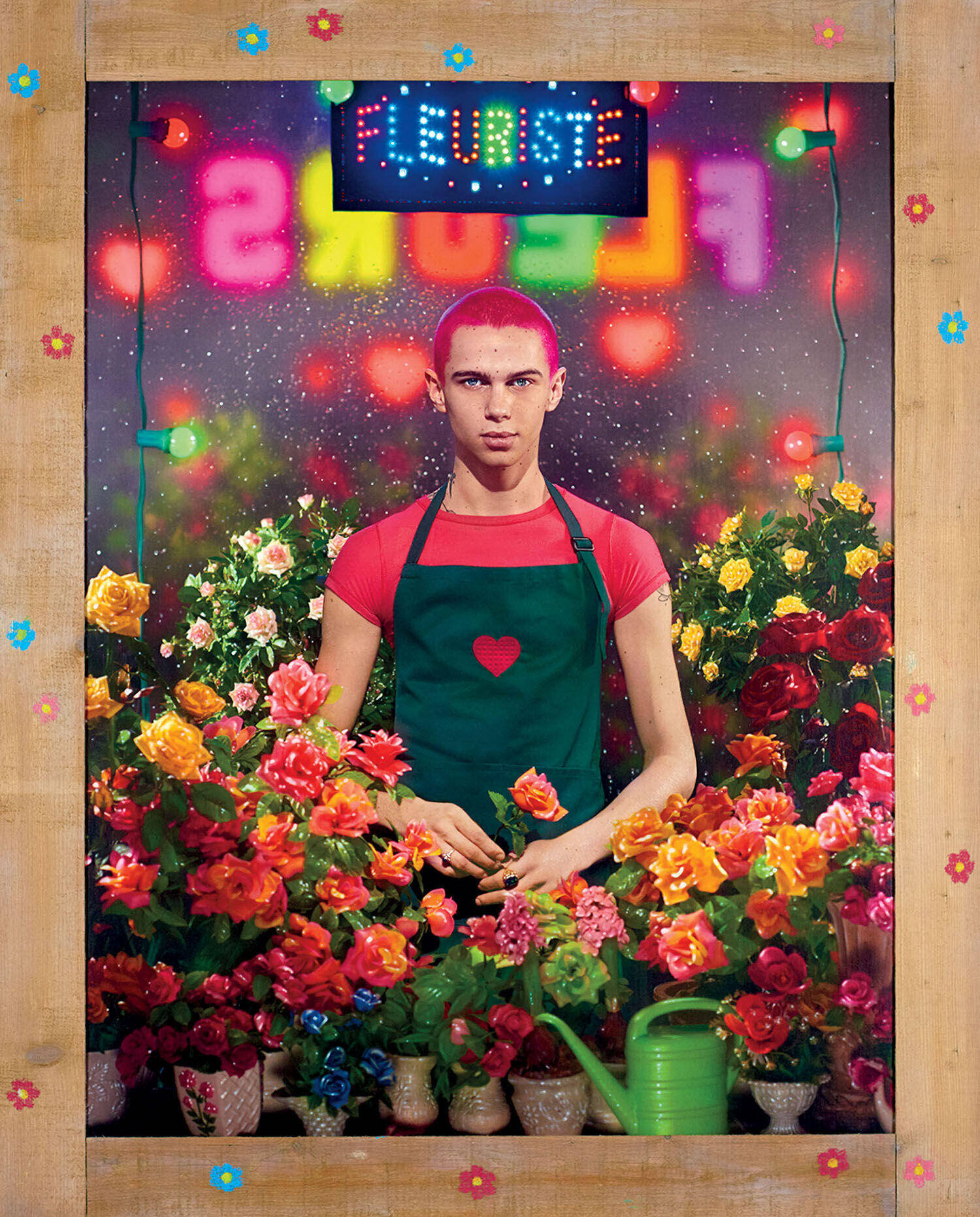A person with short pink hair wearing a green apron with a heart patch stands among many potted flowers beneath a glowing “FLEURISTE” sign.