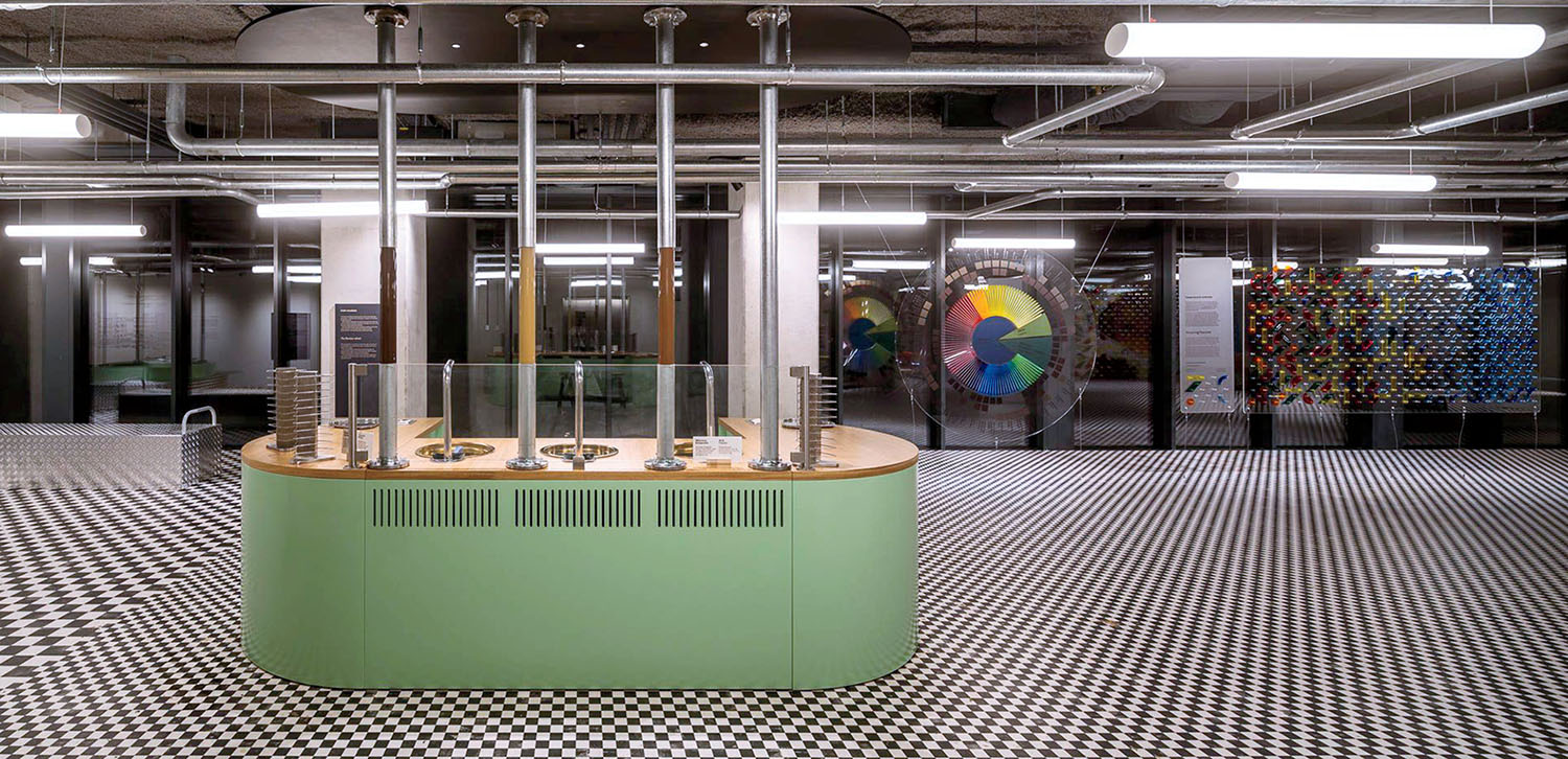 A light green, semi-circular counter with vertical pipe fixtures sits on a black-and-white checkered floor in an industrial-style space featuring color wheels and wall displays.