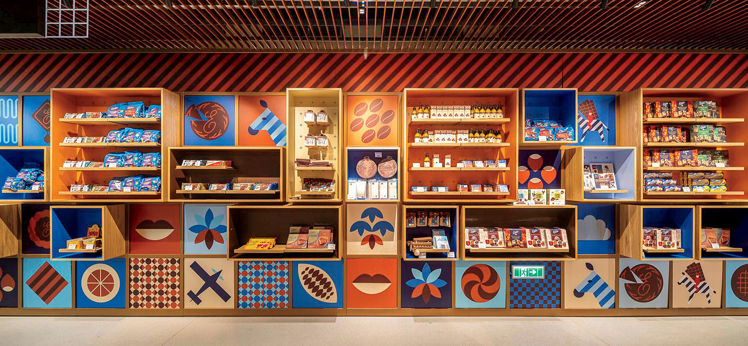 A retail wall of shelves displaying packaged chocolate bars and snacks, with decorative panels featuring geometric patterns and illustrations of cocoa beans and animals.
