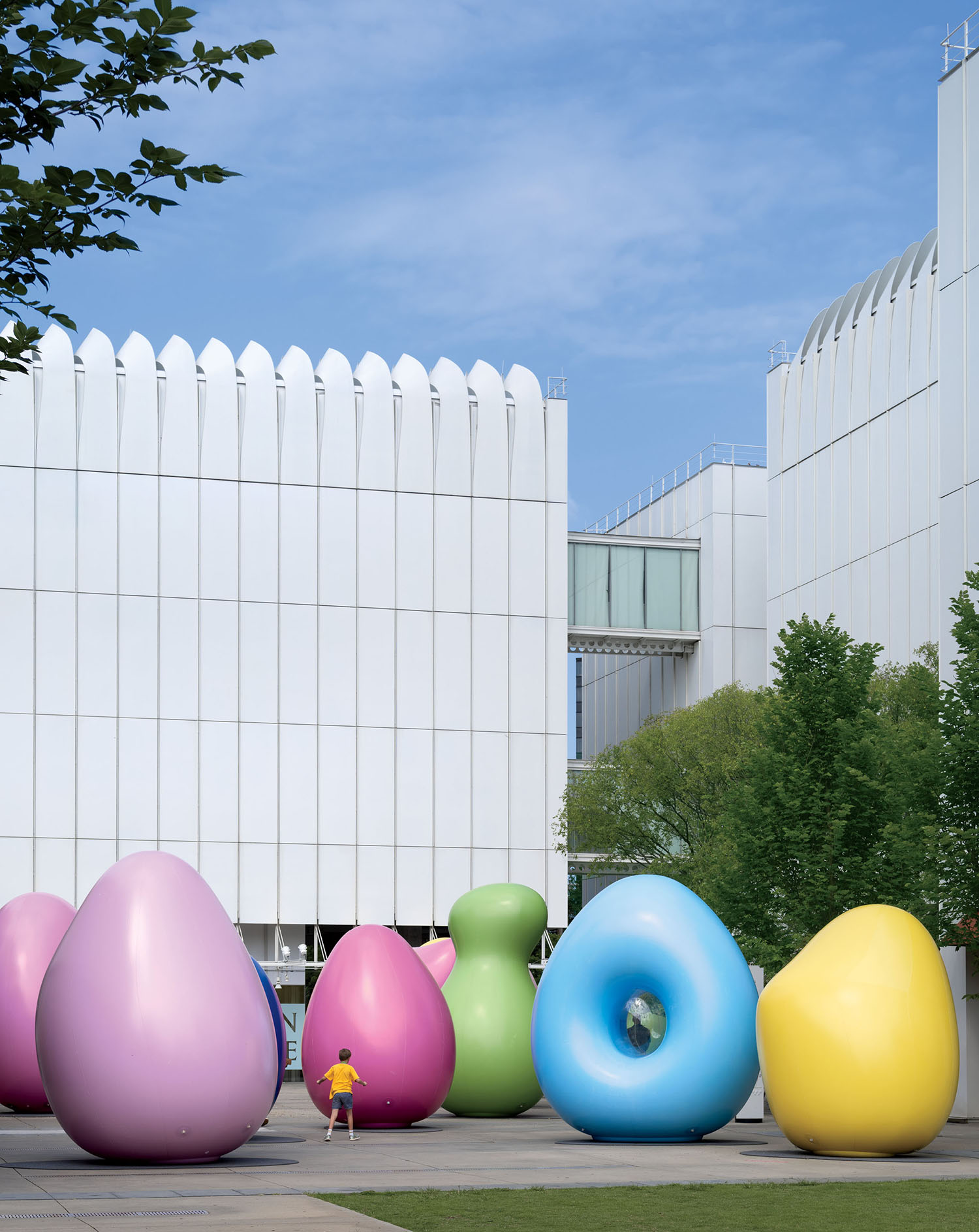 A child walks past several large, colorful, abstract sculptures in front of a modern white building.
