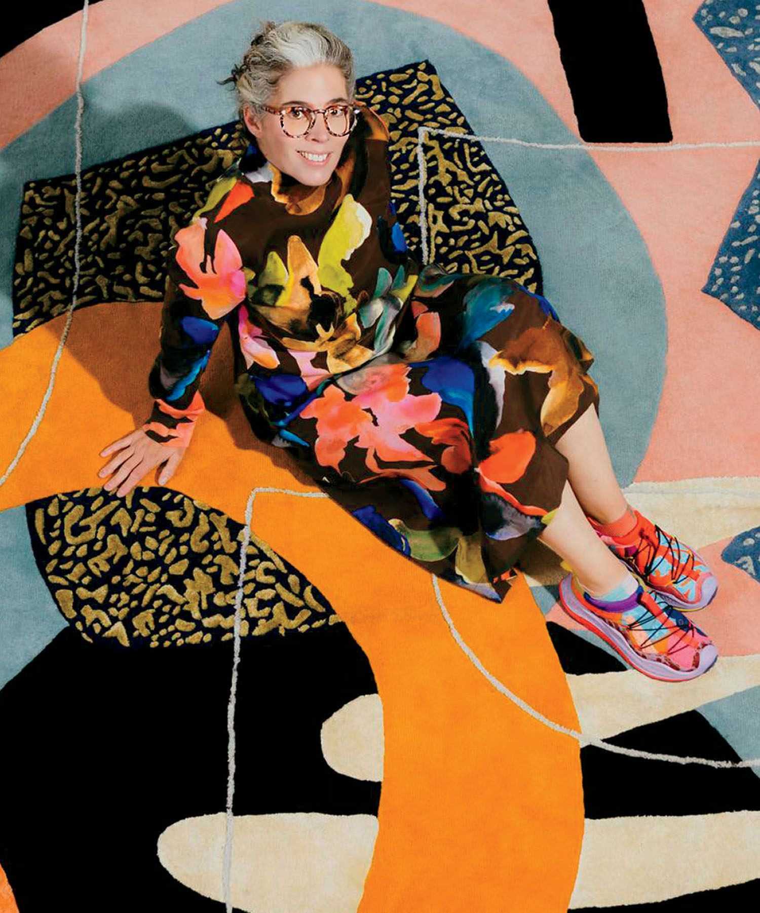A smiling woman with glasses wearing a long-sleeved floral dress and brightly colored sneakers, sitting on a large, abstract patterned rug.